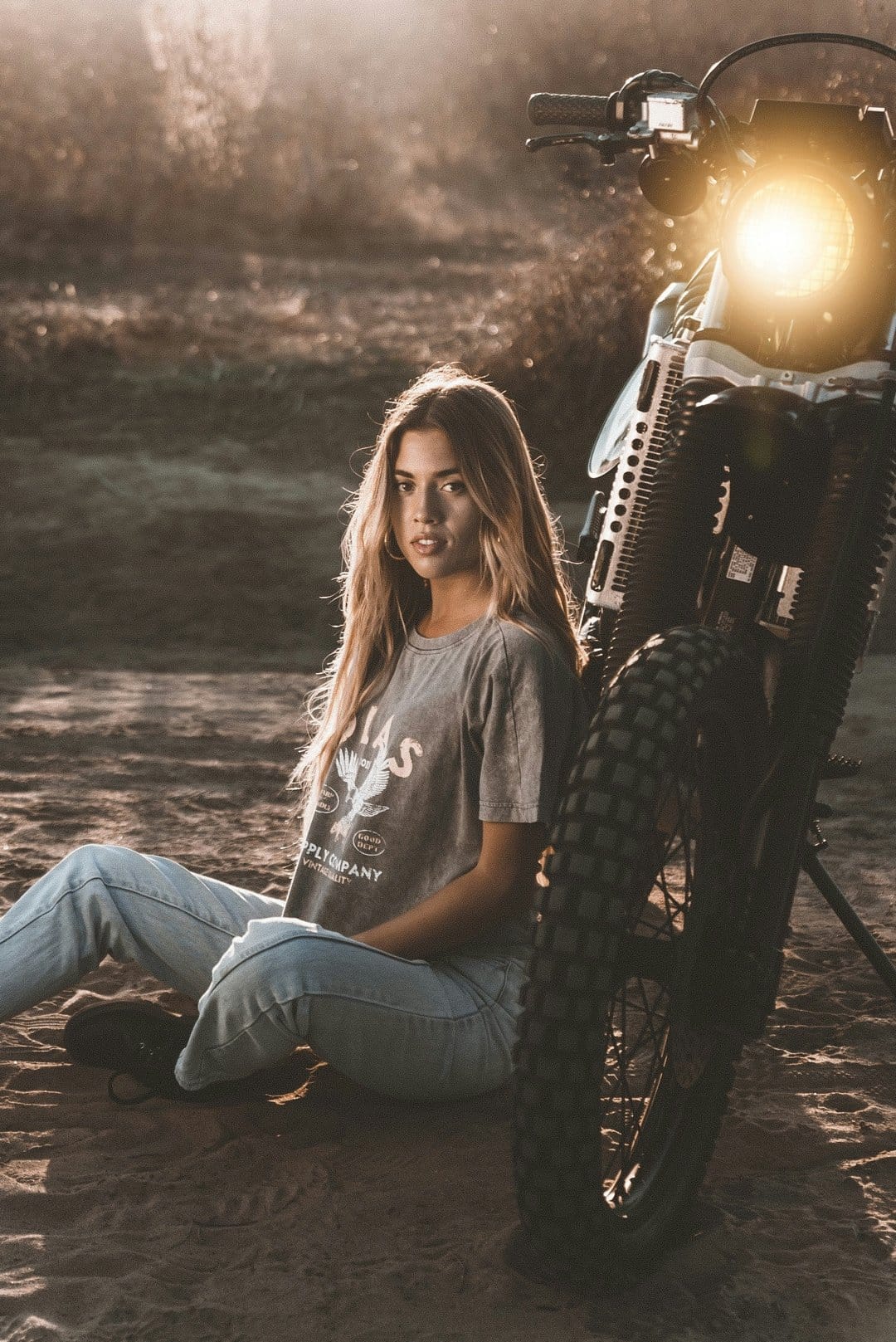Blonde woman in jeans leans against a motorcycle tire during a dusty sunset with an illuminated headlight.