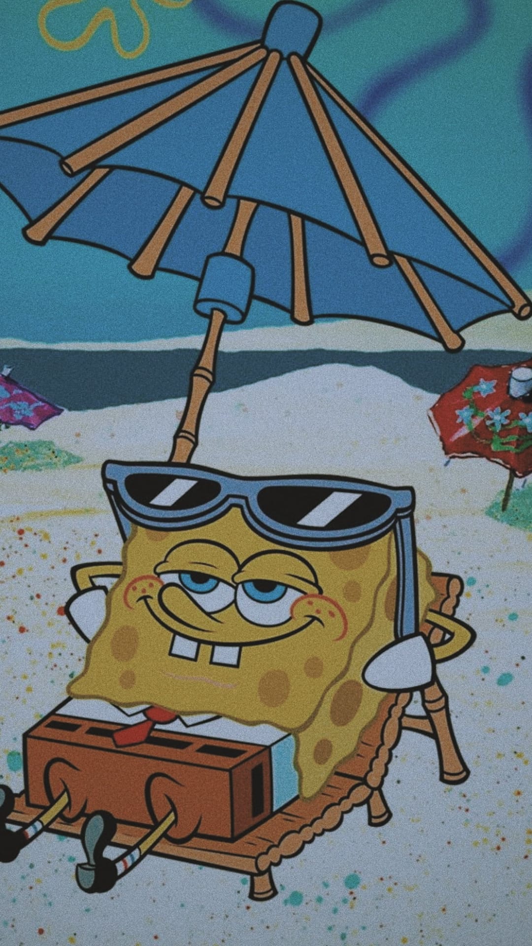 SpongeBob SquarePants wears sunglasses while lounging in a beach chair under a blue umbrella on a sandy shore.