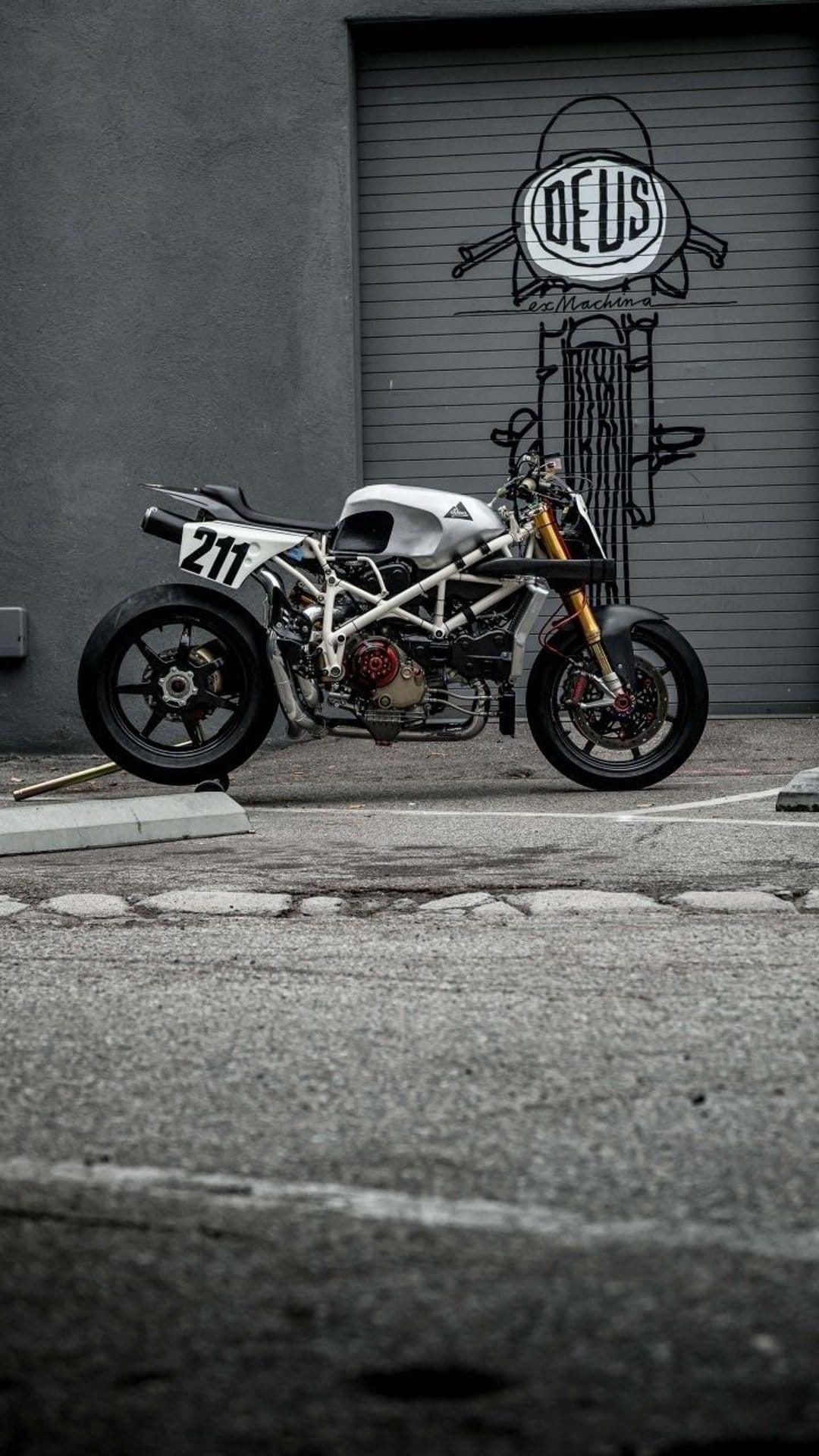 Ducati cafe racer with number 211 parked in front of a Deus Ex Machina garage door with a minimalist white frame.