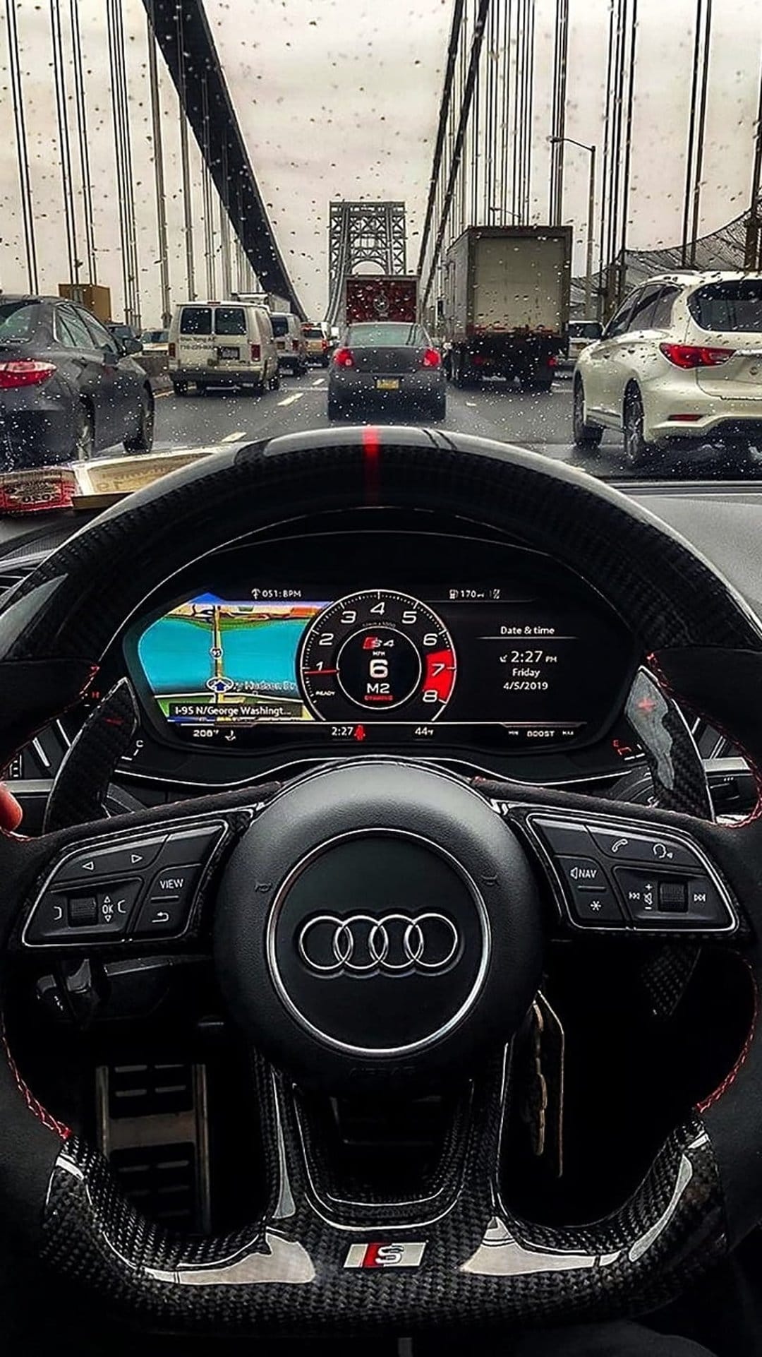 Rain-streaked car windshield looks out at a suspension bridge with heavy traffic and a glowing GPS screen.