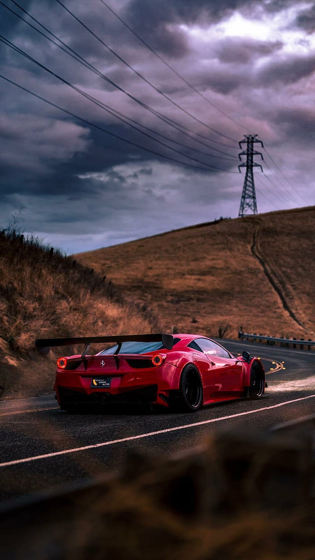 Red Ferrari supercar with a large spoiler driving on a curvy road beneath a dark, moody, overcast cloudy sky.