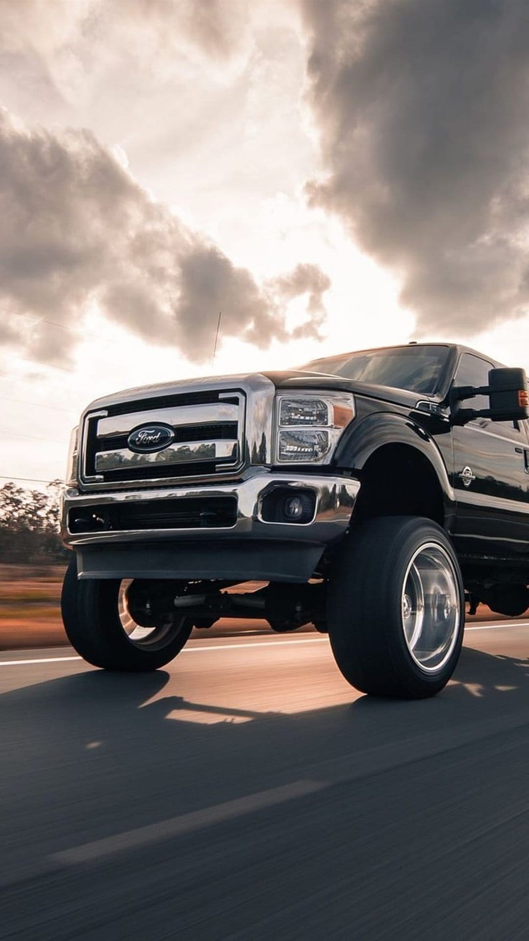 Black Ford F-Series Super Duty truck driving on an asphalt road with a chrome grille and dramatic cloudy sky.