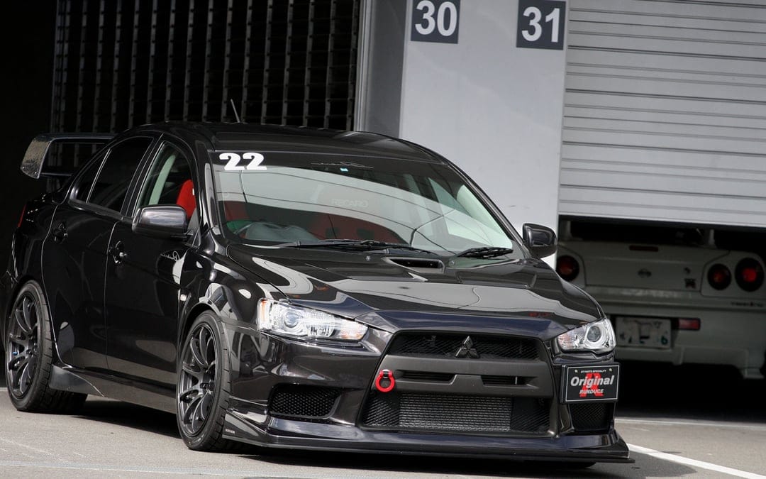 Black Mitsubishi Lancer Evolution X with red tow hook parked in a garage pit lane with a white R34 GT-R.