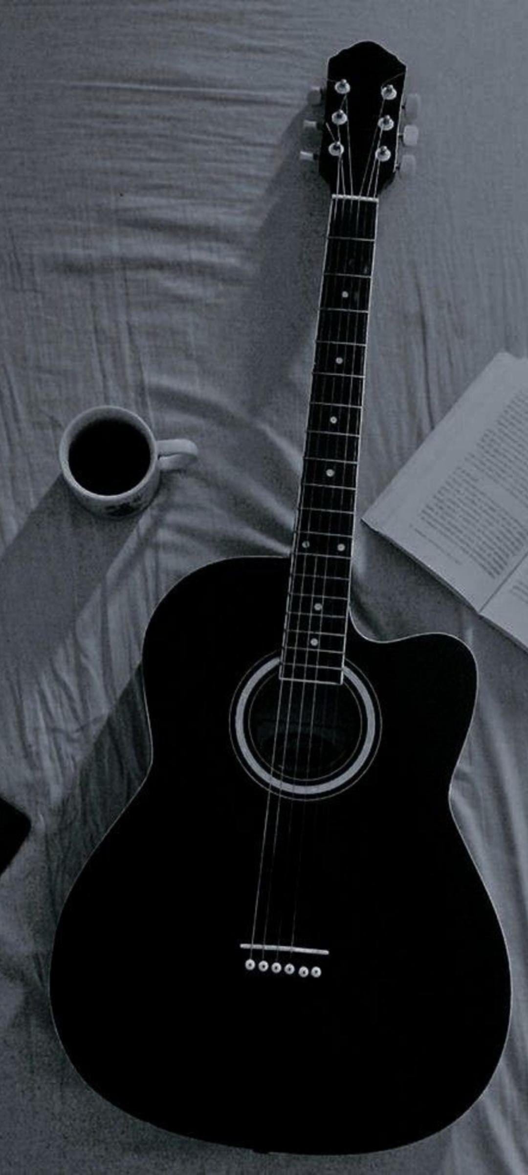 Black acoustic guitar, white ceramic mug, and an open book rest on rumpled bed sheets in a moody grayscale setting.