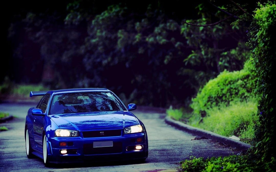 Blue Nissan Skyline R34 GTR with glowing headlights on a curved road surrounded by lush green trees.