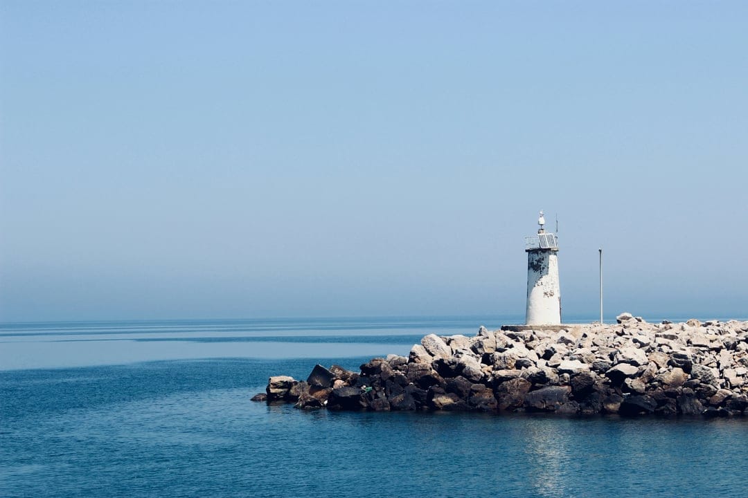 White lighthouse on a rocky jetty beside a calm blue sea under a cloudless sky during a bright day.