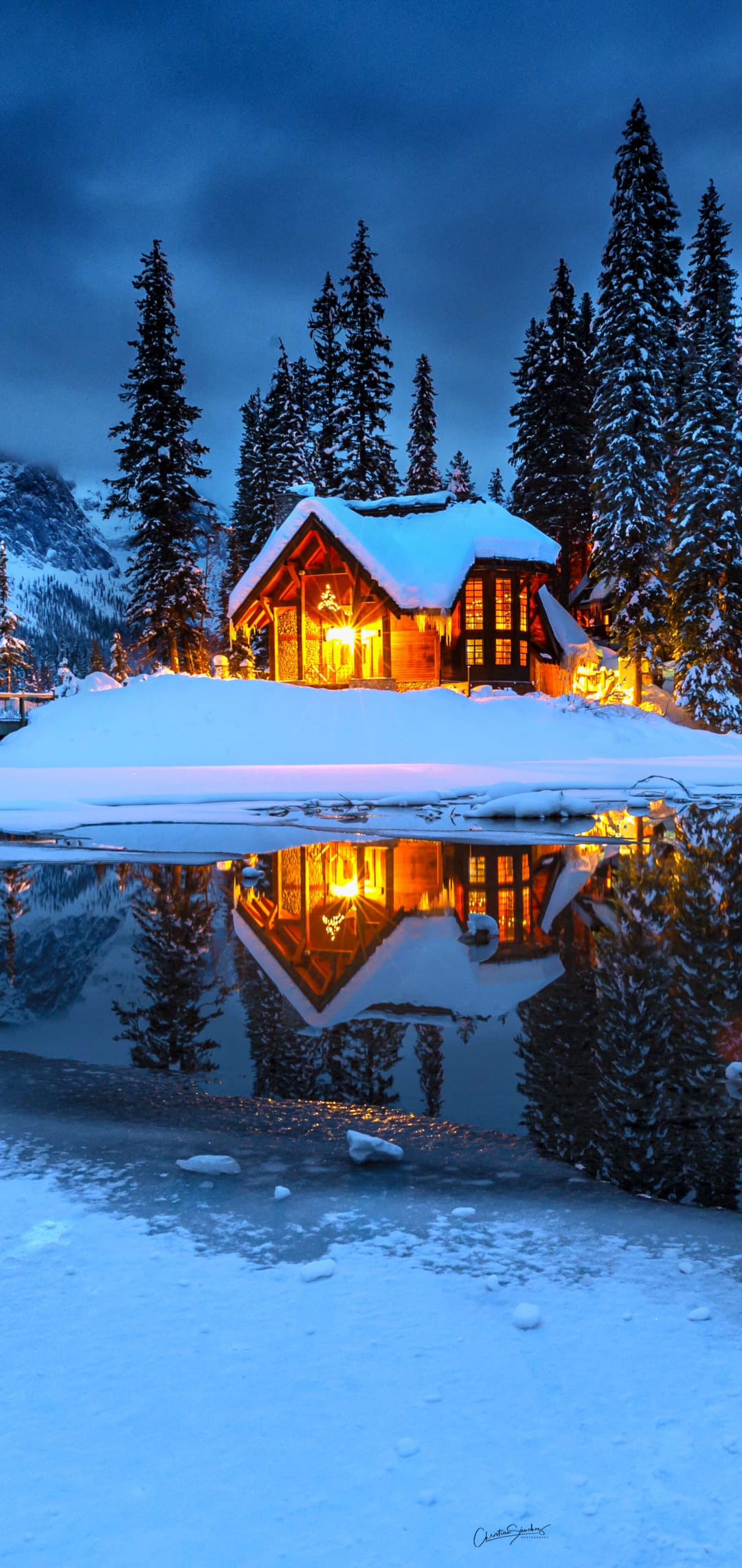Snow-dusted cabin with glowing warm lights reflects on a calm lake amidst dark winter pine trees at night.