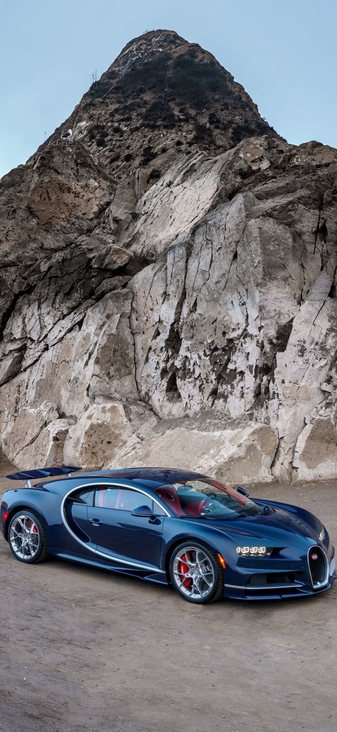 Bugatti Chiron with a red interior parked on a dirt road before a massive, textured canyon wall and desert peak.