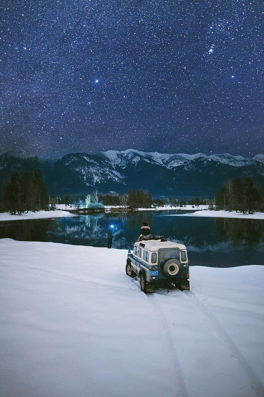 Person sitting on an off-road vehicle roof gazing at a starry night sky above snow-capped mountains and a lake.
