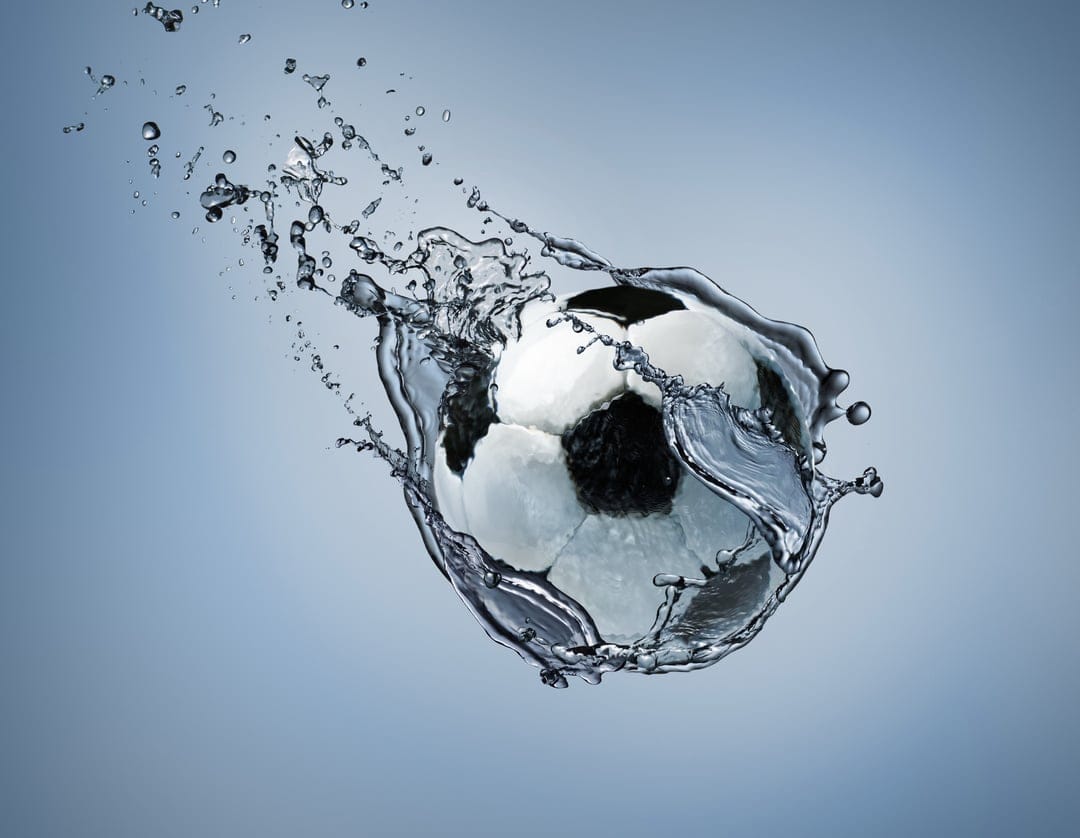 Soccer ball bursting through a splash of clear water droplets against a soft blue gradient background.