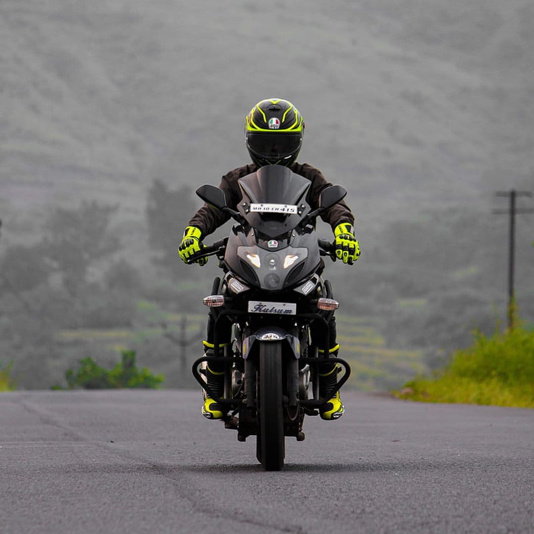 Motorcyclist in yellow gloves rides a black bike on a grey asphalt road through dense morning fog and hills.