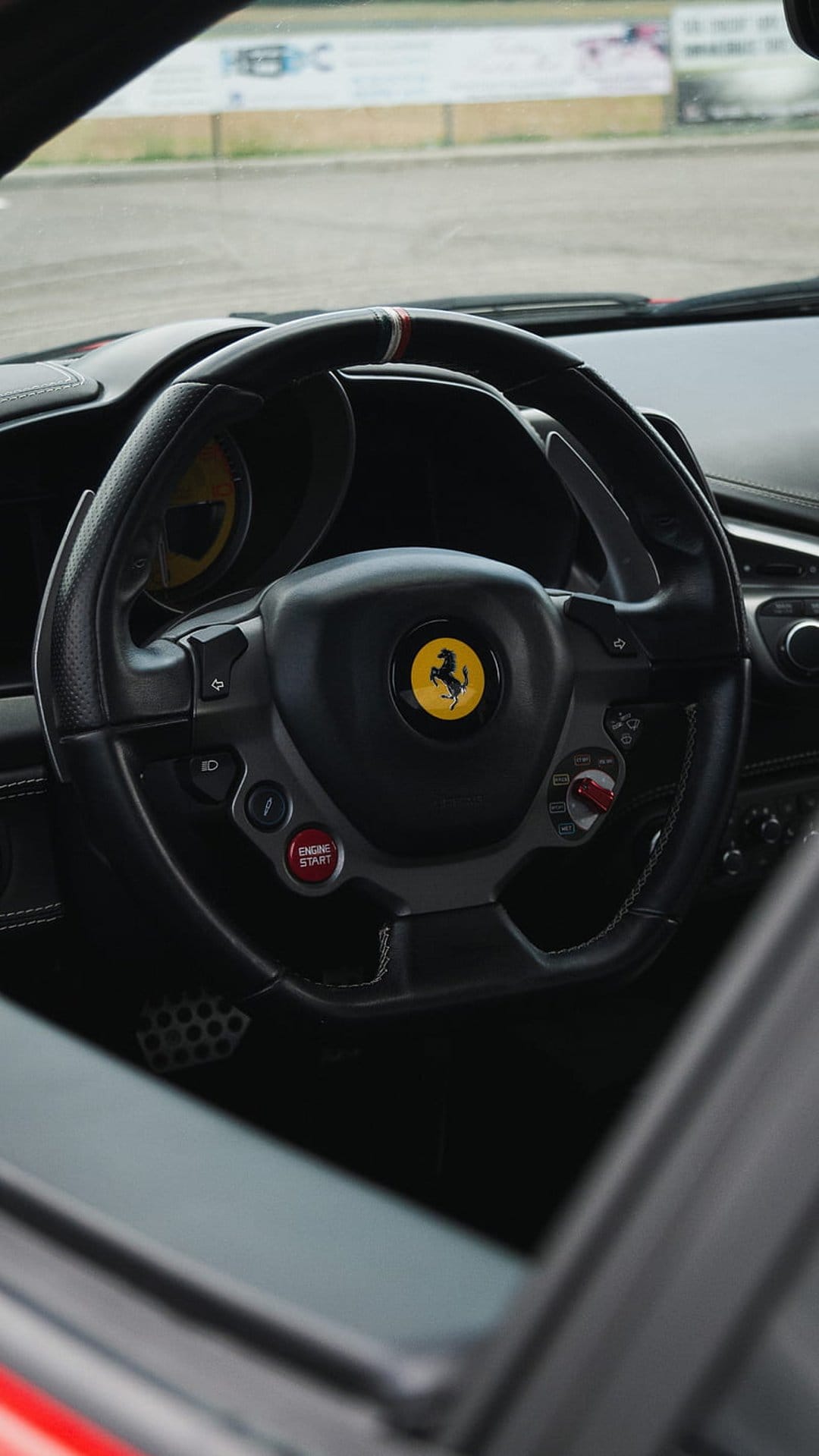 Black leather Ferrari steering wheel showcasing a yellow horse emblem, red start button, and metal paddle shifters.