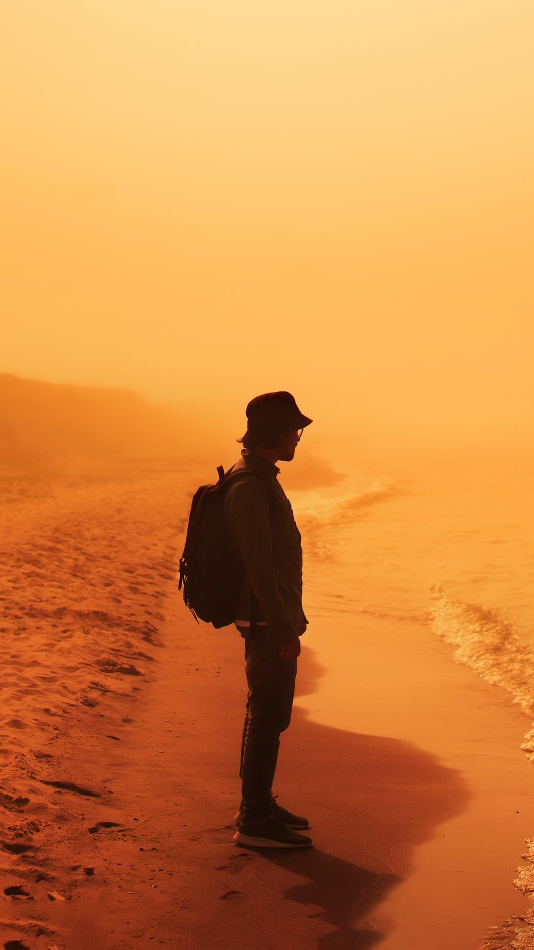 Silhouetted person with a backpack and bucket hat stands on a hazy beach during a vibrant orange coastal sunset.