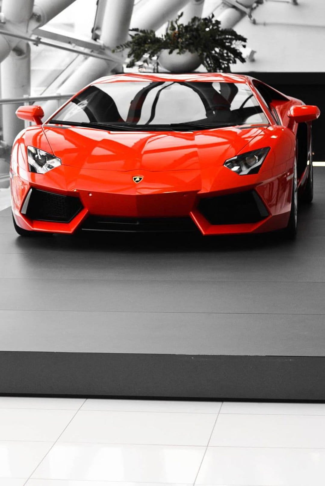 Red Lamborghini Aventador on a black stage with blurred industrial buildings and green trees in the background.