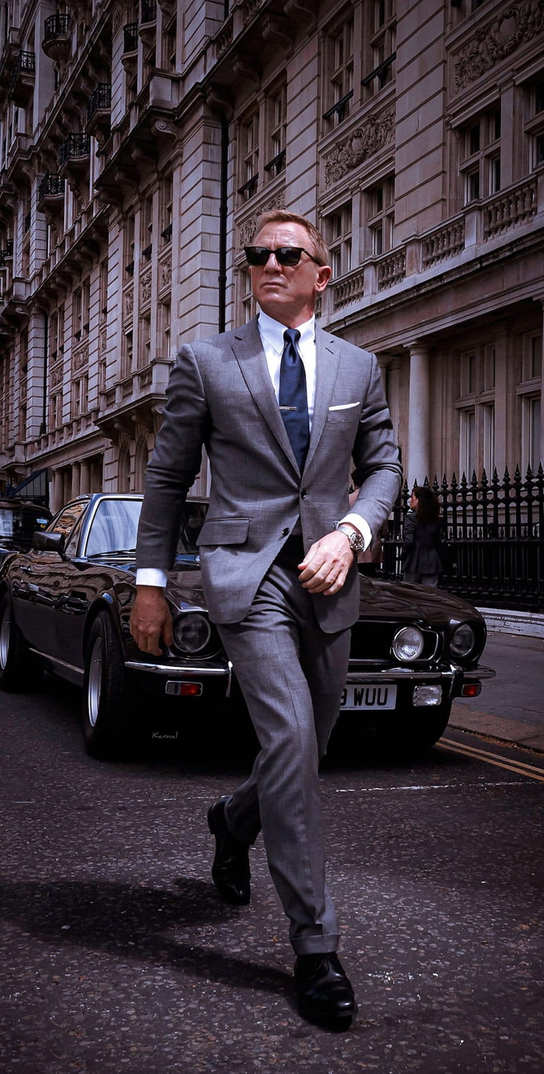 Daniel Craig as James Bond in a grey suit walks past a classic silver Aston Martin on a London street.