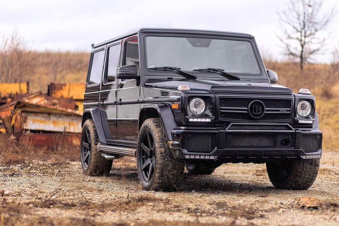 Black Brabus G-Class SUV with muddy tires on a dirt path against a backdrop of rusted industrial metal.