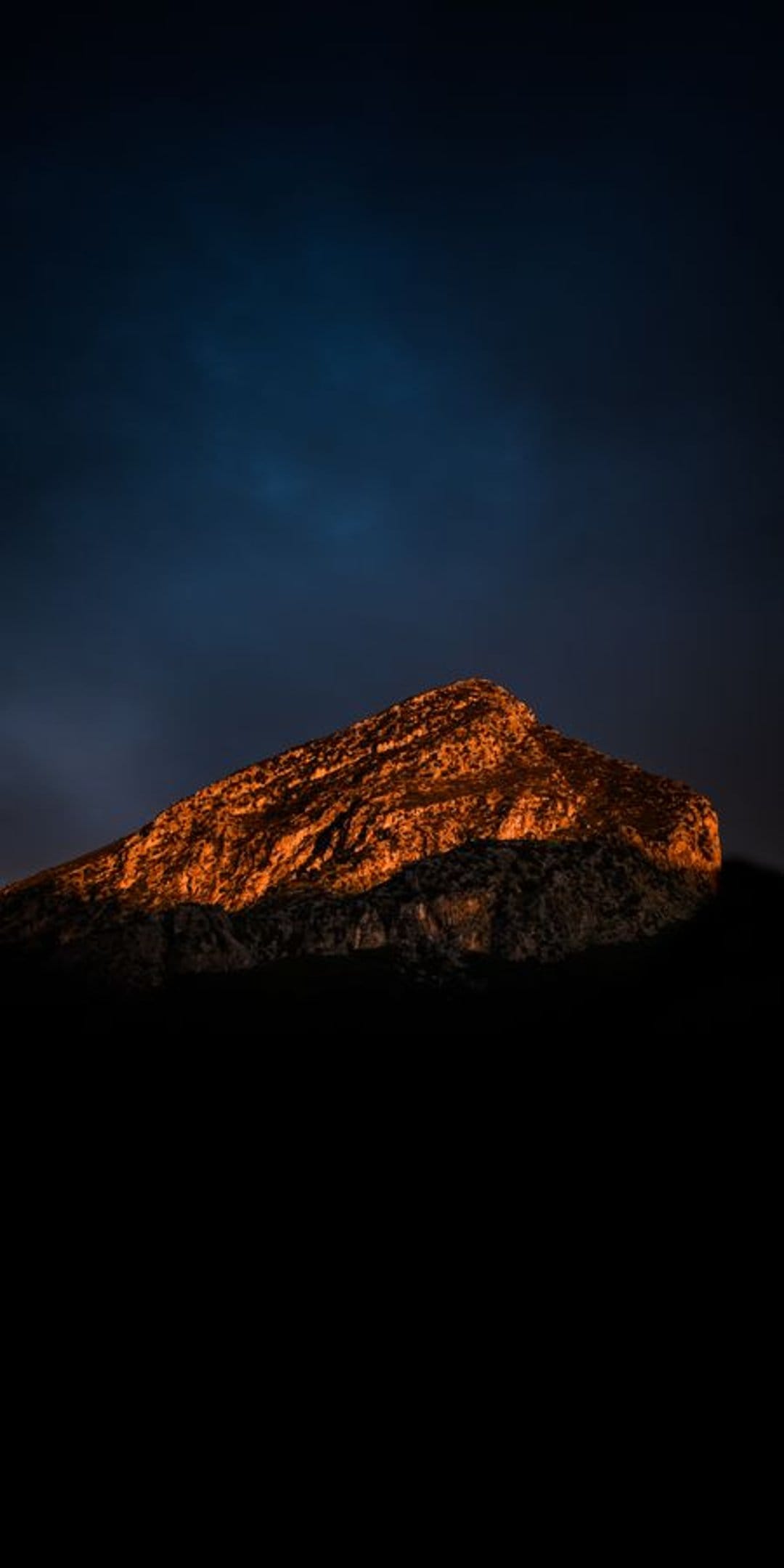 Jagged mountain summit glowing with golden light against a dark blue sky with shadowed rock in the foreground.