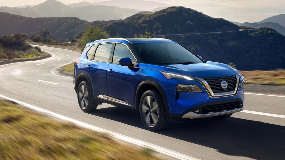 Blue Nissan Rogue SUV driving on a winding asphalt road with green hills and mountains in the background.