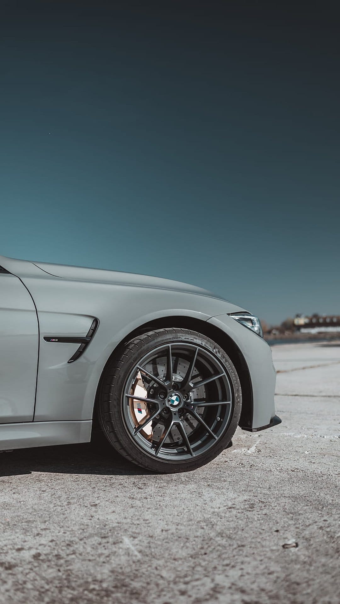 BMW M4 alloy wheel and brake caliper resting on textured concrete pavement against a vivid blue sky backdrop.