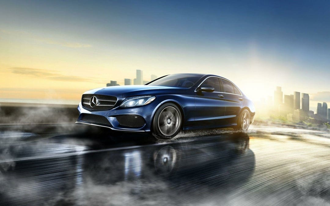 Blue Mercedes-Benz sedan driving on a wet road with motion blur and a city skyline at sunset in the background.