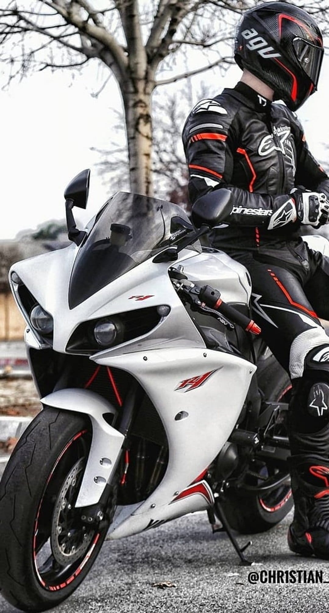 Motorcyclist in red and black racing gear sits on a white Yamaha R1 sportbike on asphalt near a bare tree.