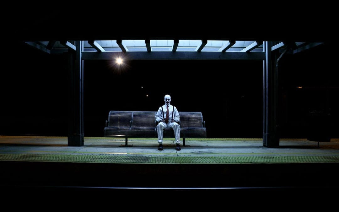 Man in clown makeup wearing a suit sits on a bench under glowing bus stop lights against a black night sky.