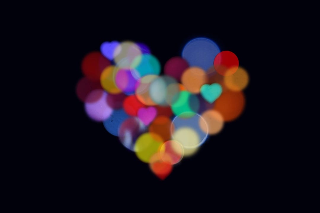 Heart-shaped arrangement of colorful out-of-focus bokeh lights shimmering against a pitch-black background.