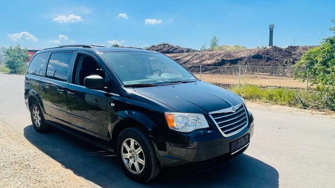 Black Chrysler minivan parked on a sunlit road with dirt mounds and a clear blue sky in the background.