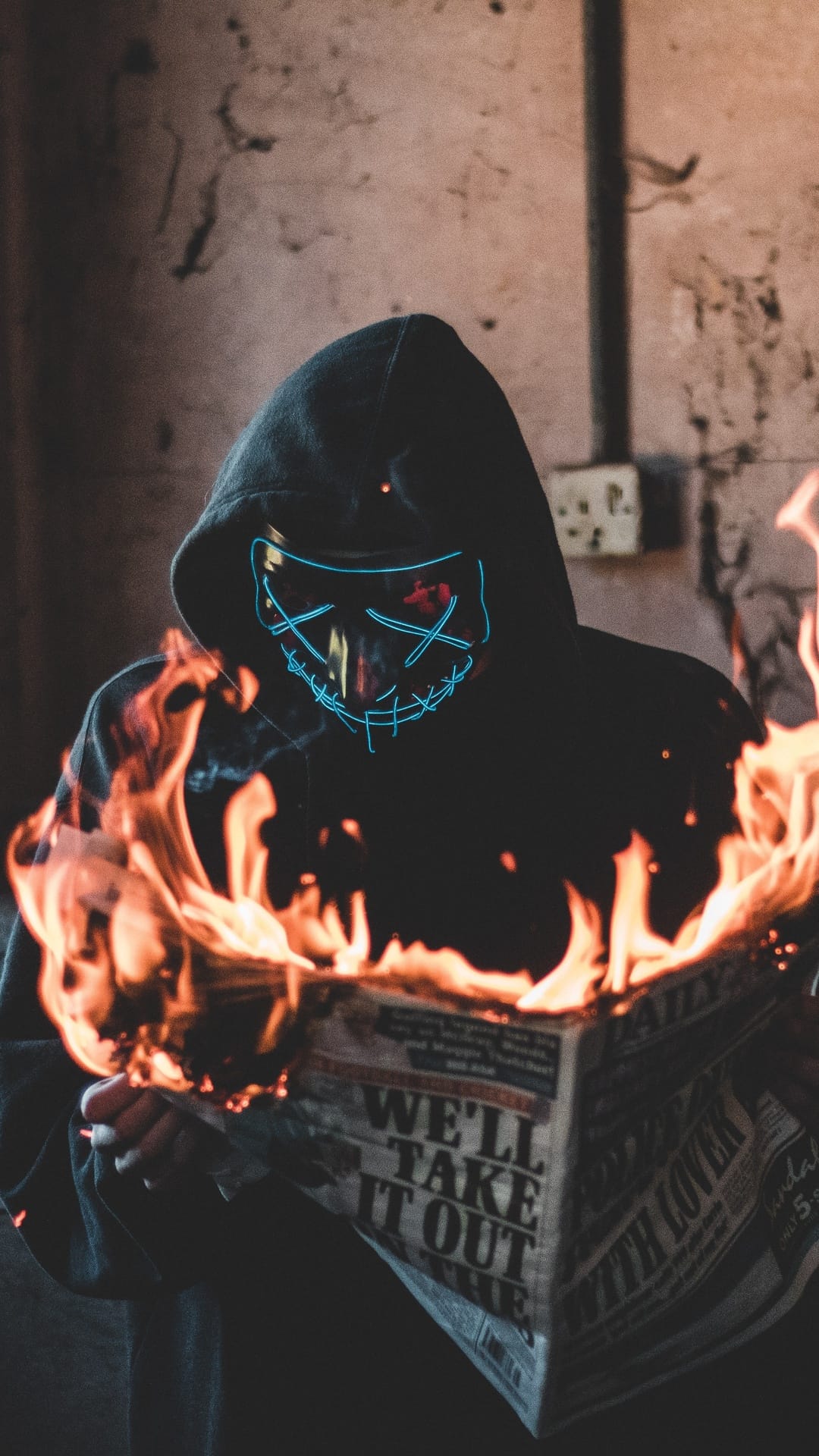 Masked person in a dark hoodie holds a burning newspaper with glowing blue X eyes in an industrial setting.