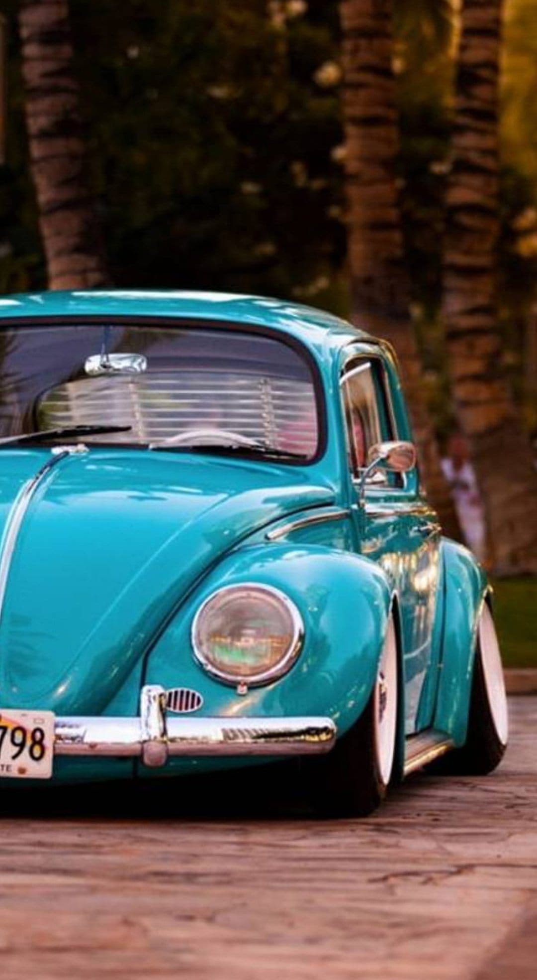Turquoise Volkswagen Beetle with a chrome bumper parked on wooden planks under sunset light and palm trees.