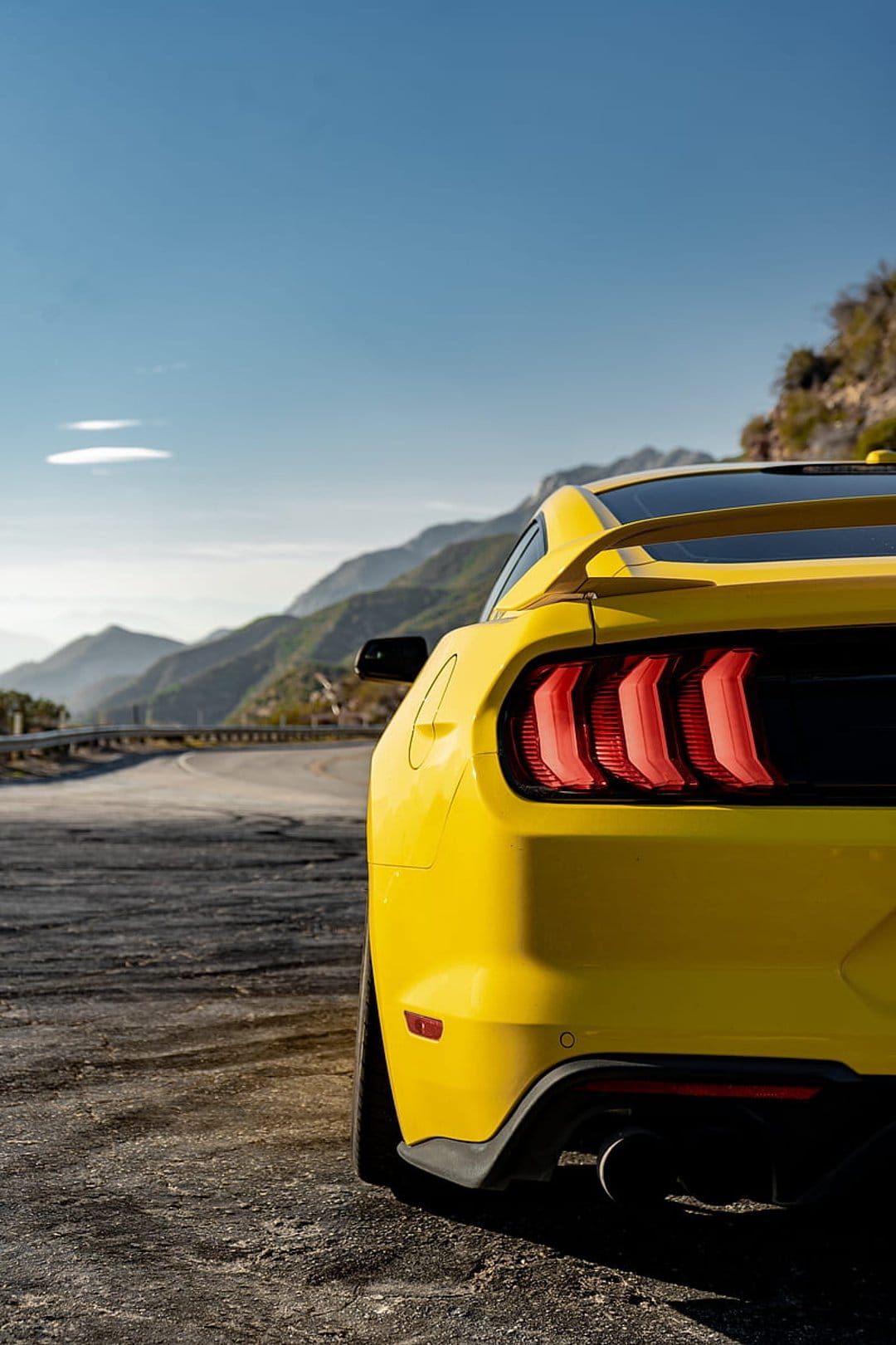 Yellow Ford Mustang rear with glowing red taillights parked on a winding mountain road under a clear blue sky.