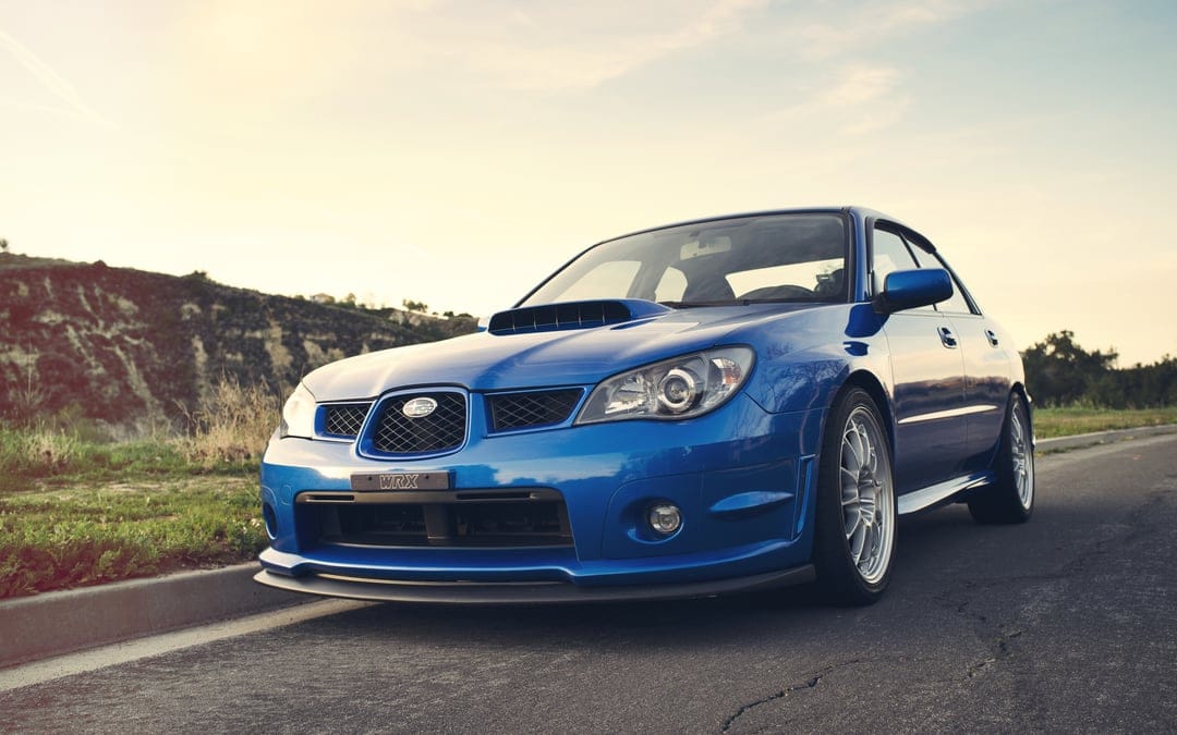 Blue Subaru WRX sedan parked on a paved road at sunset with silver wheels and a visible hood scoop.