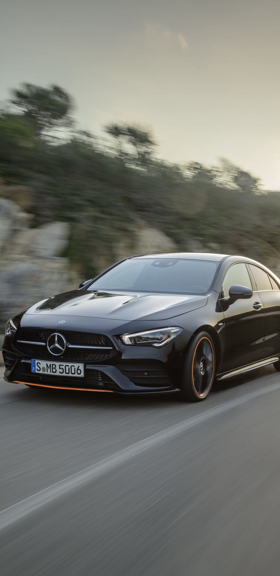 Black Mercedes-Benz CLA with orange trim driving on a road with motion blur and rocky hills background.