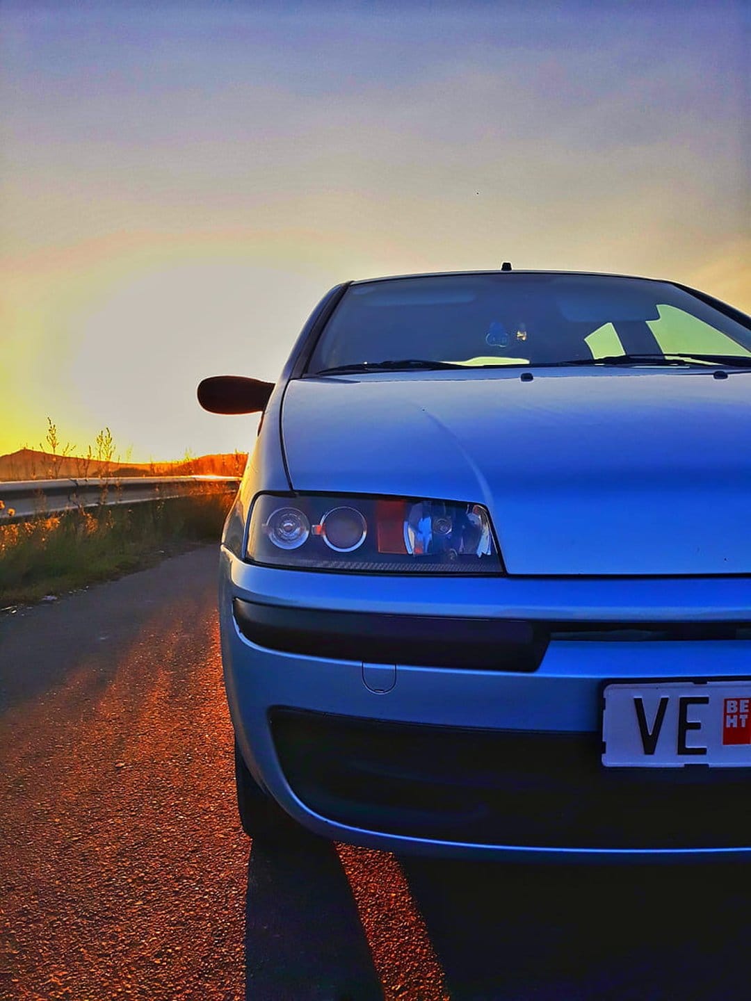 Blue Fiat Punto parked on a rural road at sunset with golden light hitting the front grille and headlights.