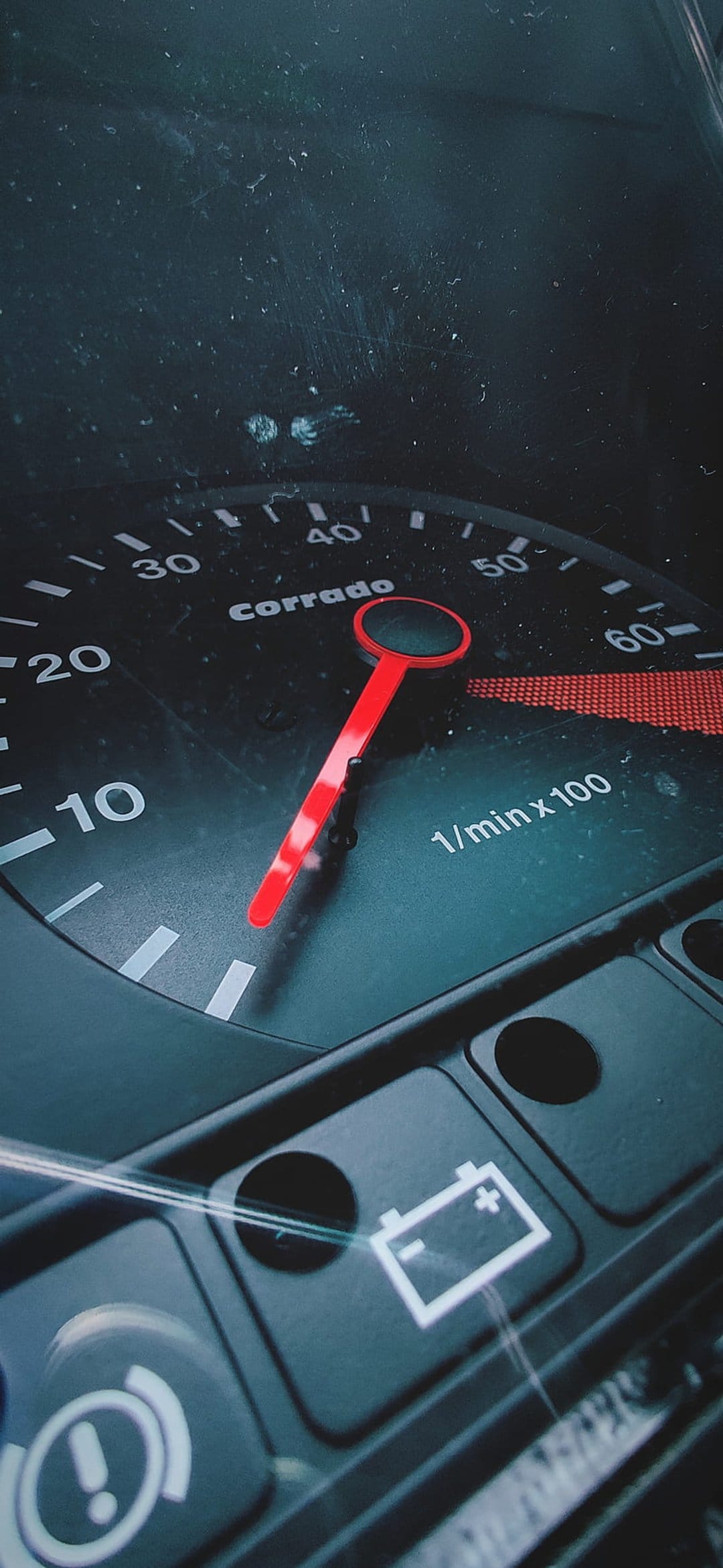 Analog car tachometer with Corrado branding, RPM needle, and glowing red battery warning light on a dusty lens.
