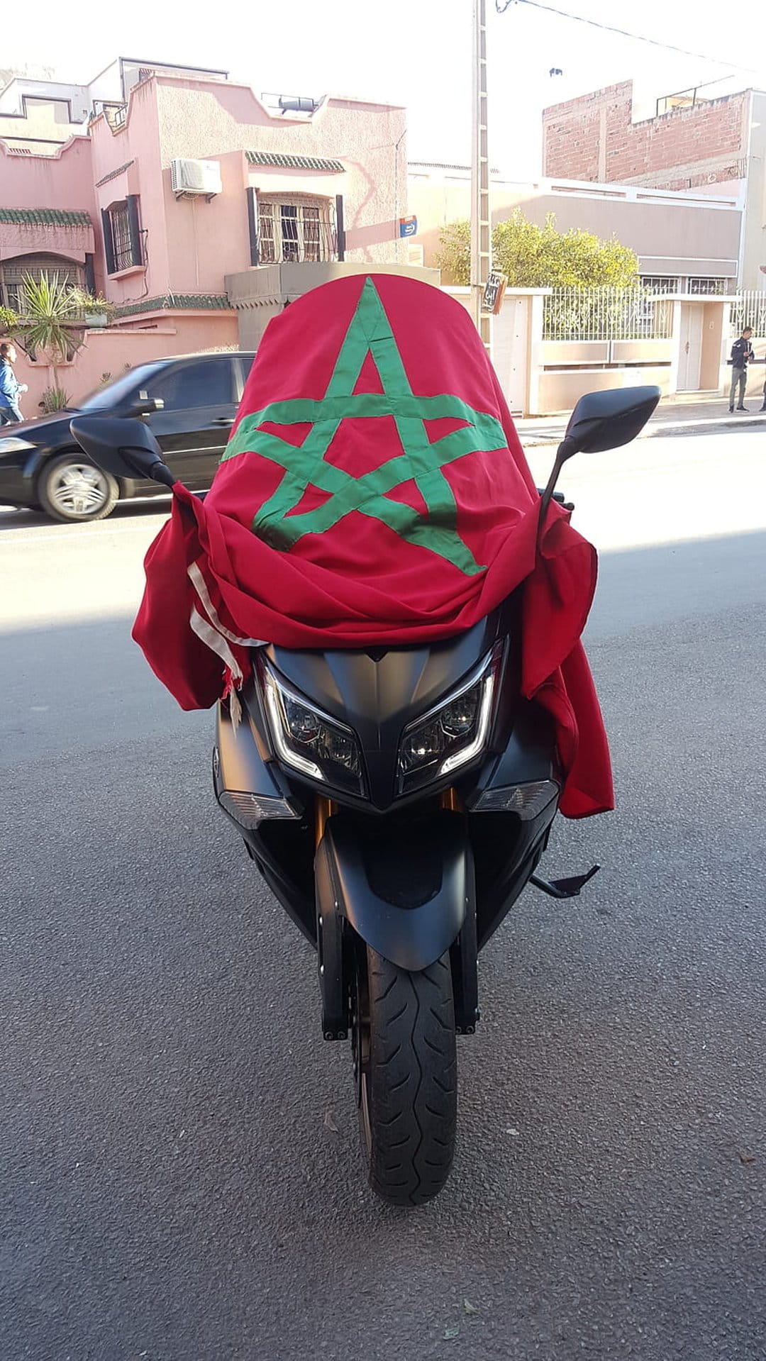 Moroccan flag with a green star draped over a black motor scooter parked on an asphalt city street.