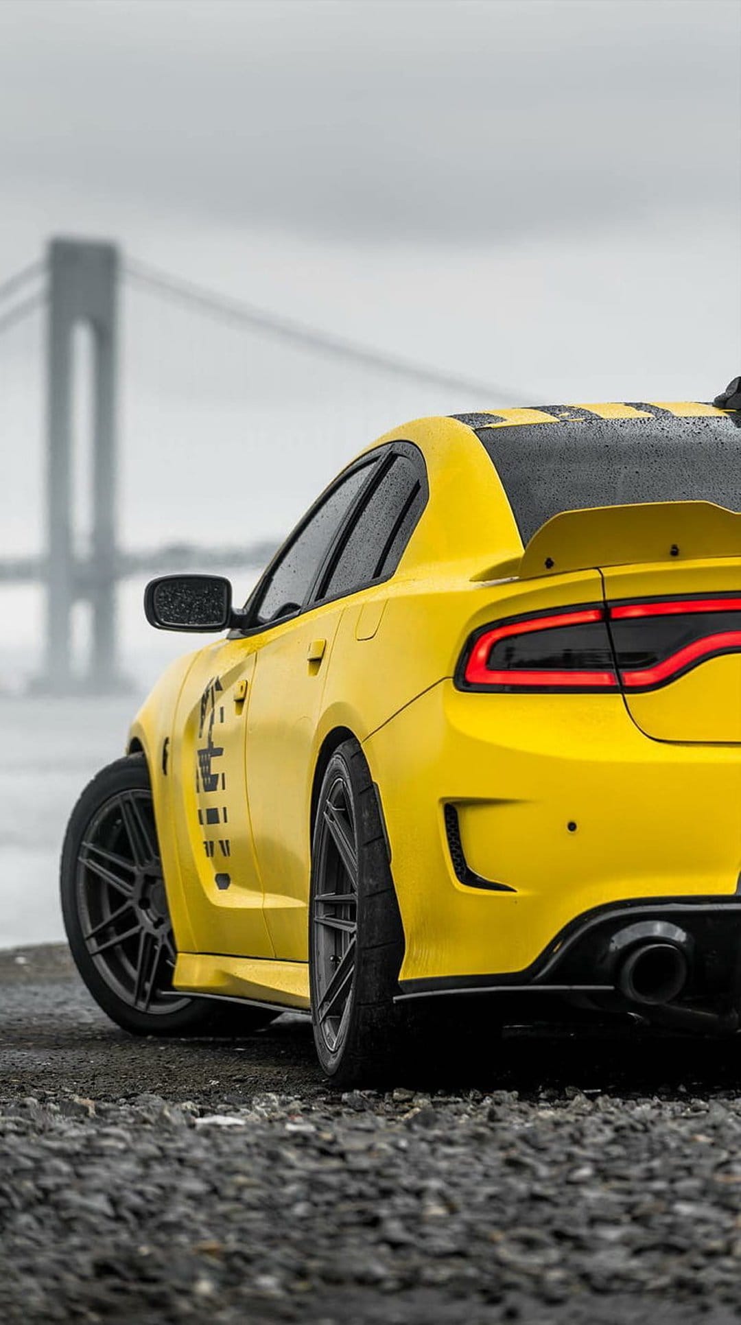 Yellow Dodge Charger with black stripes parked on wet gravel near a bridge during a rainy day with lit taillights.