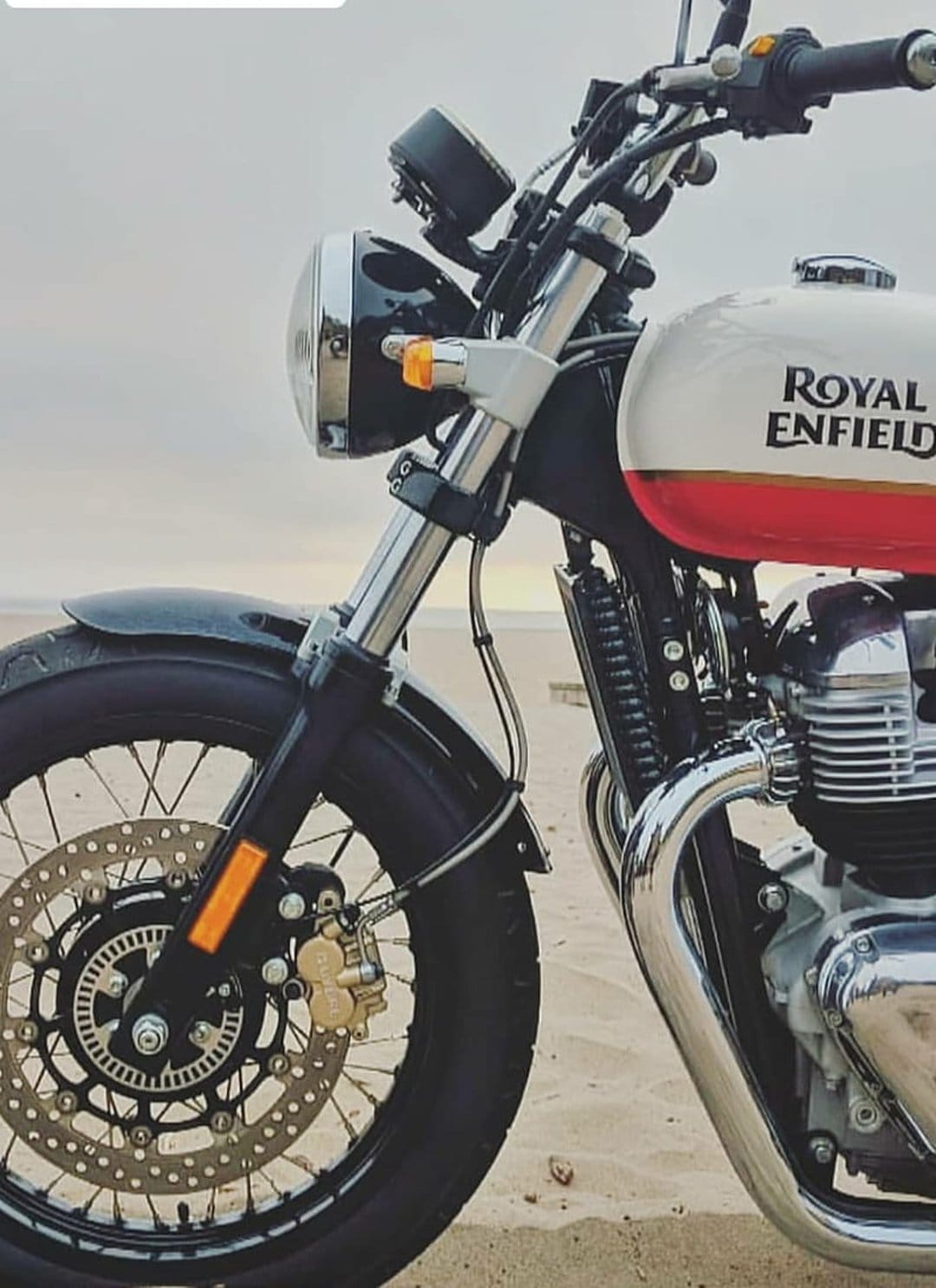 Royal Enfield motorcycle with chrome accents on a sandy beach under a cloudy sky with ocean waves in the distance.