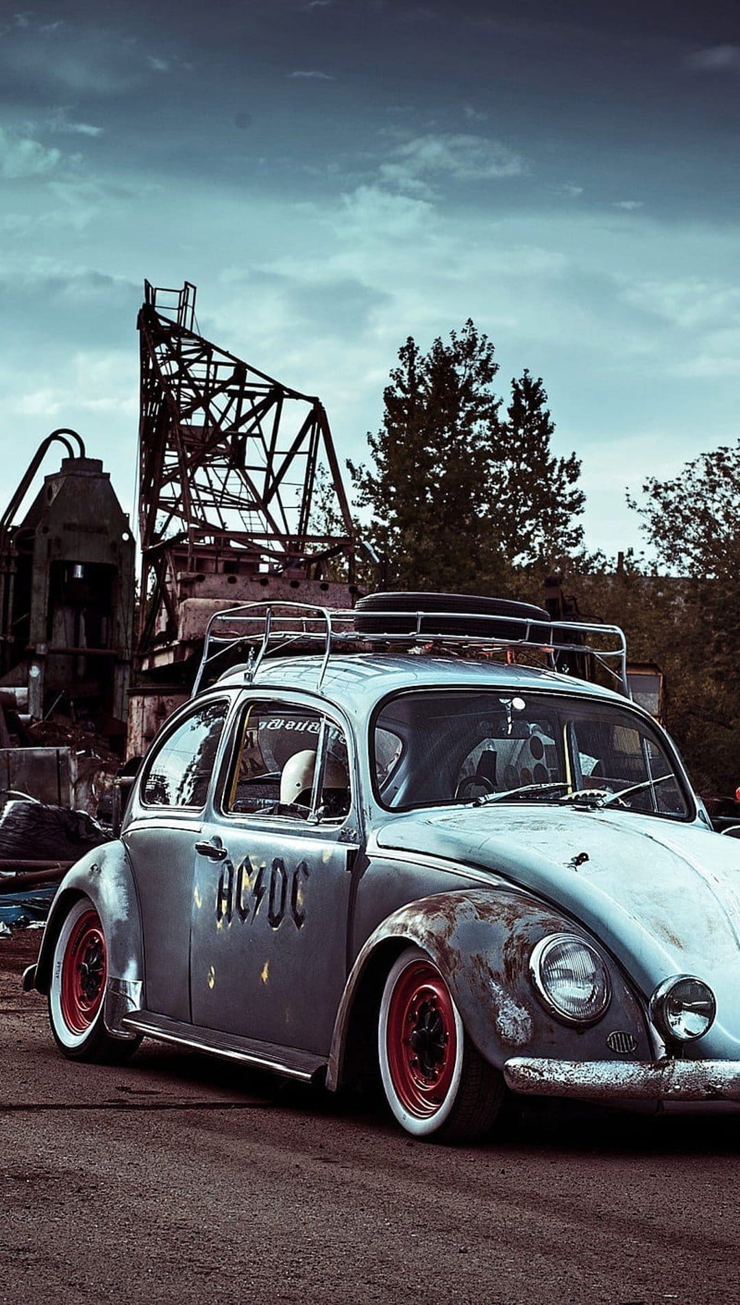 Rusty Volkswagen Beetle with AC/DC lettering parked near industrial gears under a moody, overcast sky.