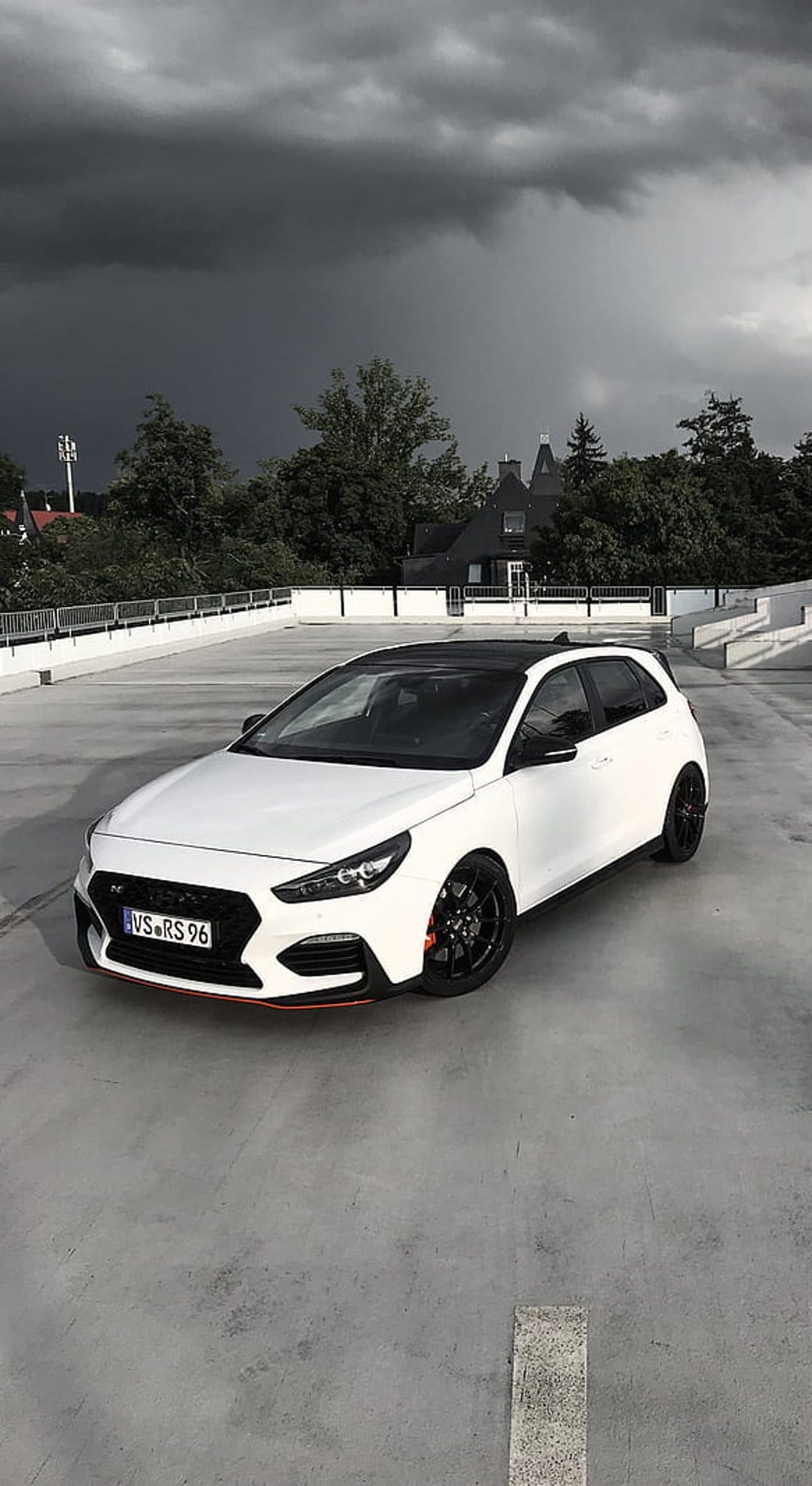 White Hyundai i30 N with red trim parked on concrete under a dark stormy sky near green trees and a building.