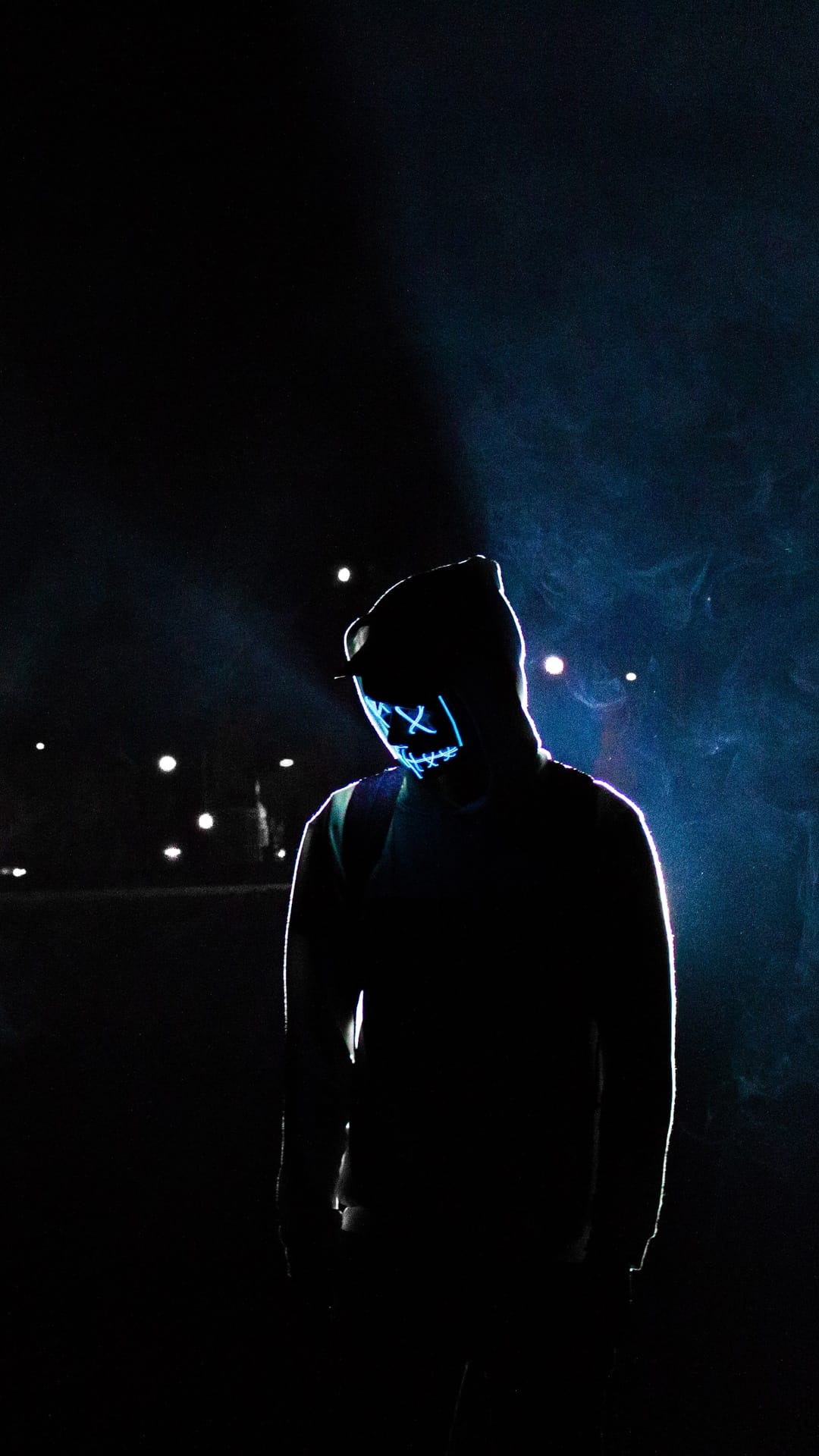 Person in a glowing blue neon mask with X-shaped eyes and a stitched mouth standing in a smoky night setting.