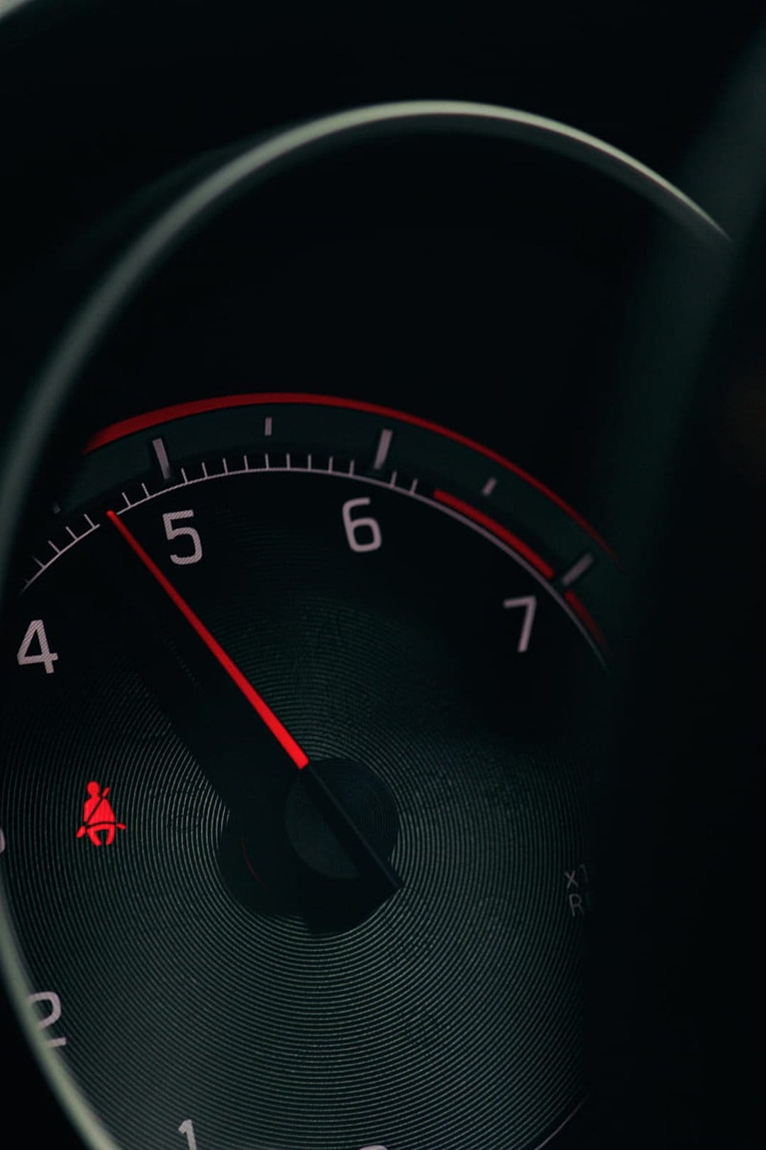 Red tachometer needle pointing to five thousand RPM beside a glowing seatbelt warning light on a dark dashboard.