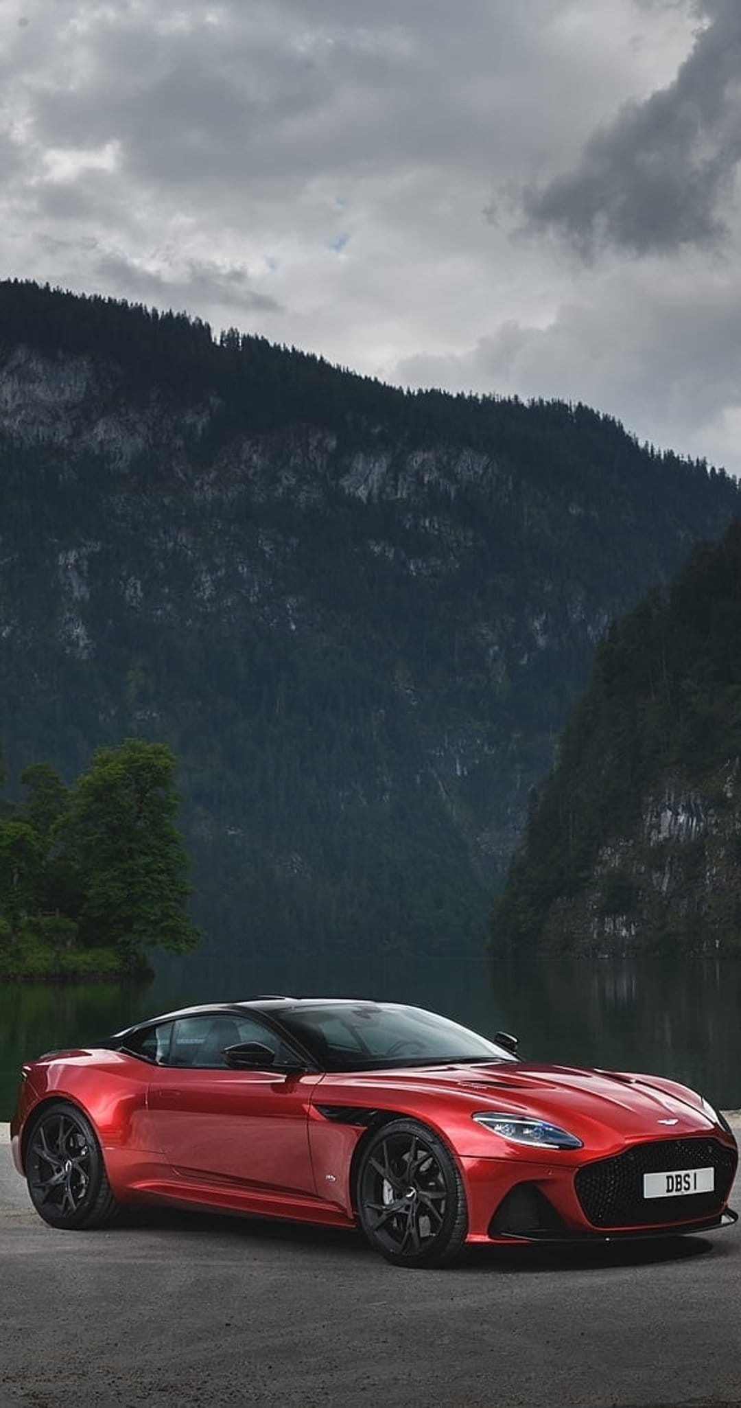 Red Aston Martin DBS Superleggera parked on asphalt near a mountain lake under an overcast cloudy sky.