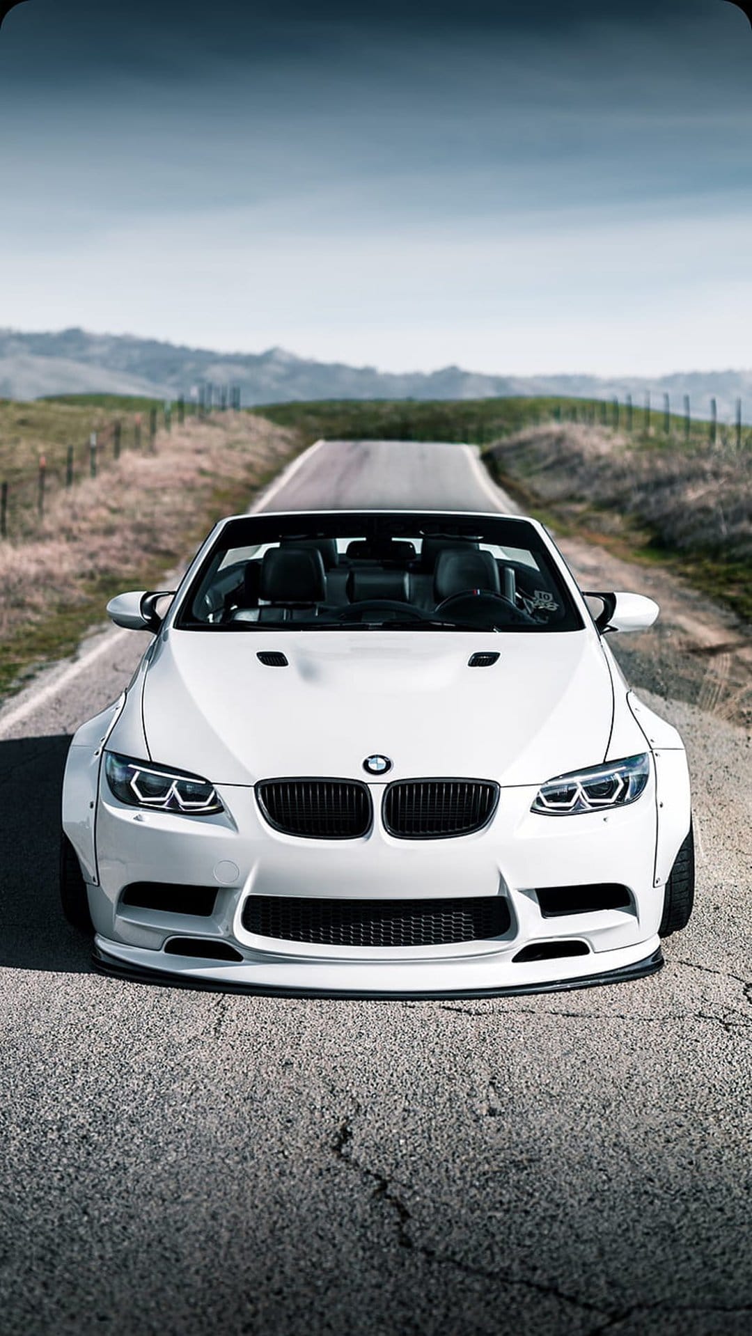White BMW convertible with a widebody kit parked on a cracked asphalt road leading into rolling green hills.