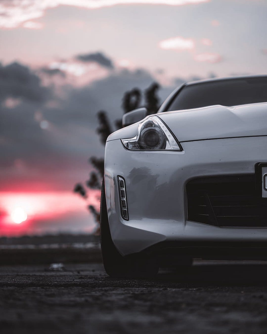 White sports car front grille and glowing headlights set against a dramatic pink and orange cloudy sunset sky.