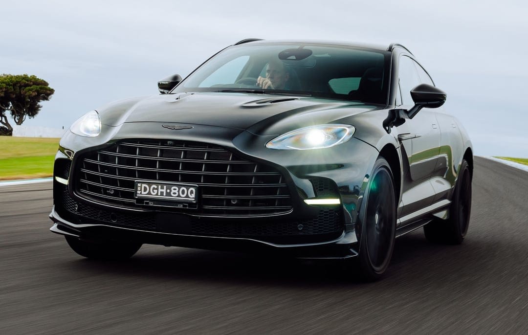 Deep green Aston Martin DBX707 luxury SUV with lit headlights speeding on a paved racetrack circuit.