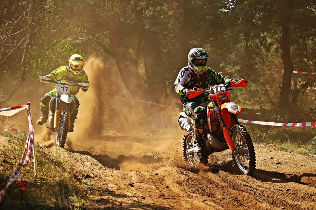Two dirt bike racers kick up brown dust while speeding past red and white tape on a wooded motocross track.