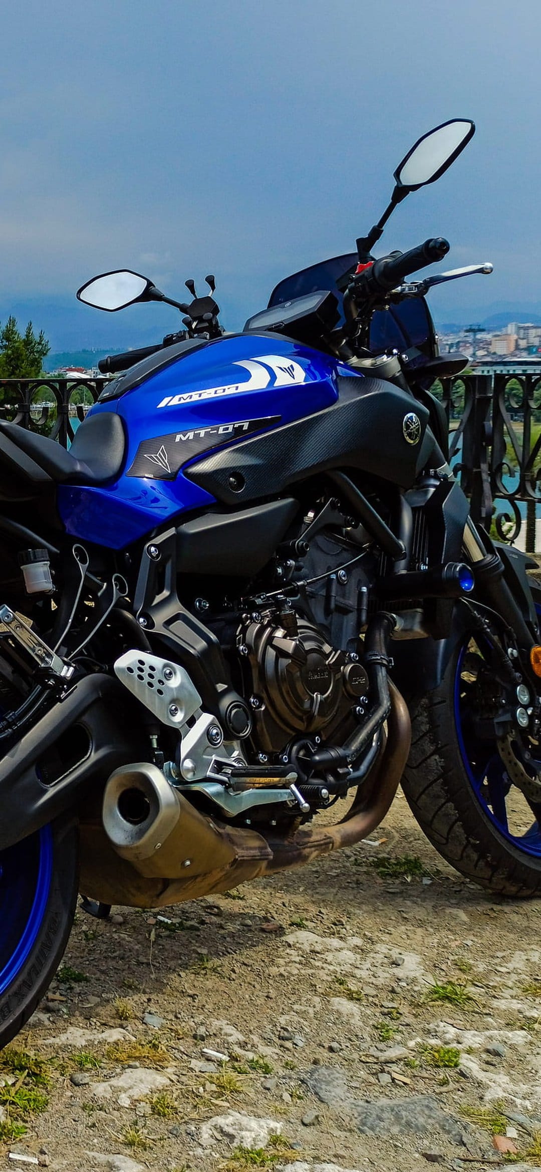 Yamaha MT-07 motorcycle with a blue fuel tank parked on a rocky surface under a cloudy sky with a city view.