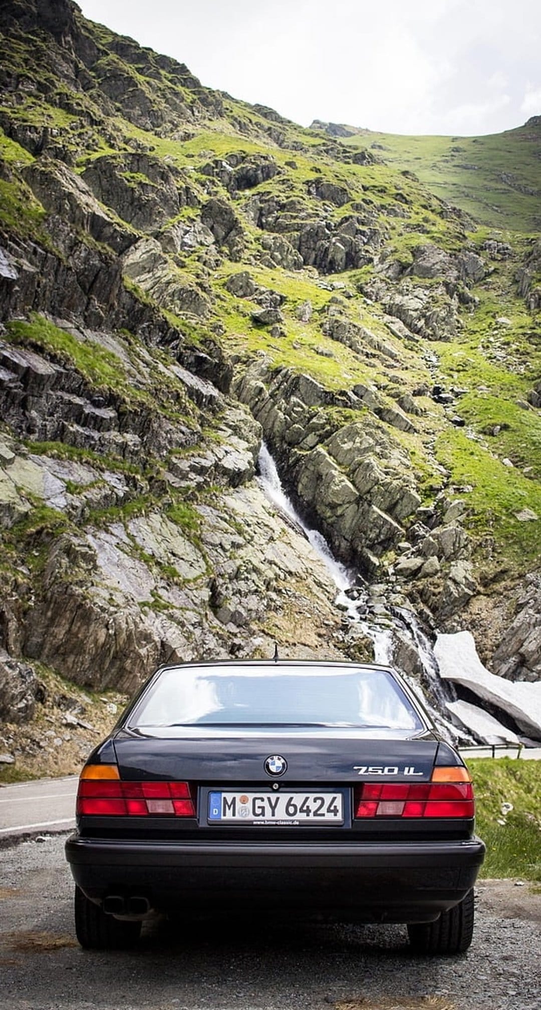 Black BMW 750iL luxury sedan parked on a winding asphalt road next to a tall cascading mountain waterfall.