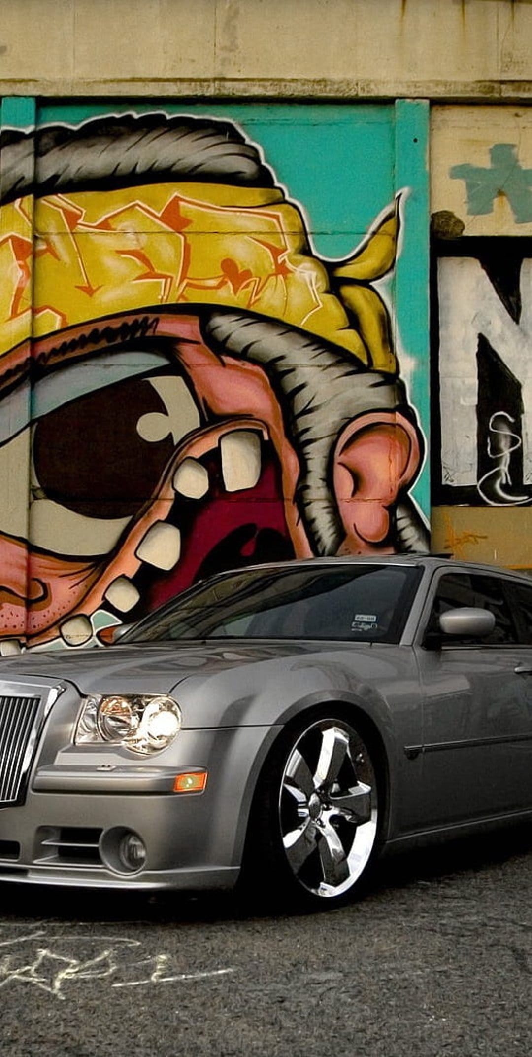 Chrysler 300 sedan in silver parked before a vibrant cartoon face graffiti mural in a gritty city alleyway.